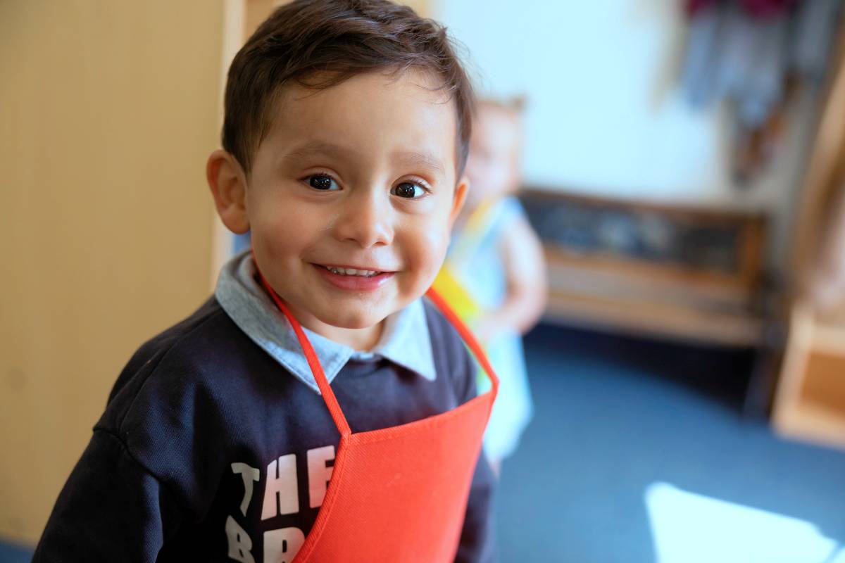 SNACS pre-k student wearing a bright red apron, looking at the camera and smiling.